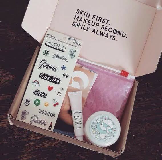 inside of a glossier gift box. there are beauty products and stickers. inside the top of the box has the text ‘skin first. makeup second. smile always.’