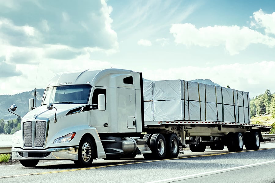 White semi truck hauling a tarped flatbed trailer along a scenic mountain highway